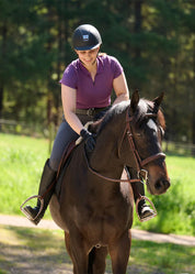 Front view of a female model wearing the purple Kerrits Serenity Coolcore Short Sleeve Polo while riding a dark colored horse
