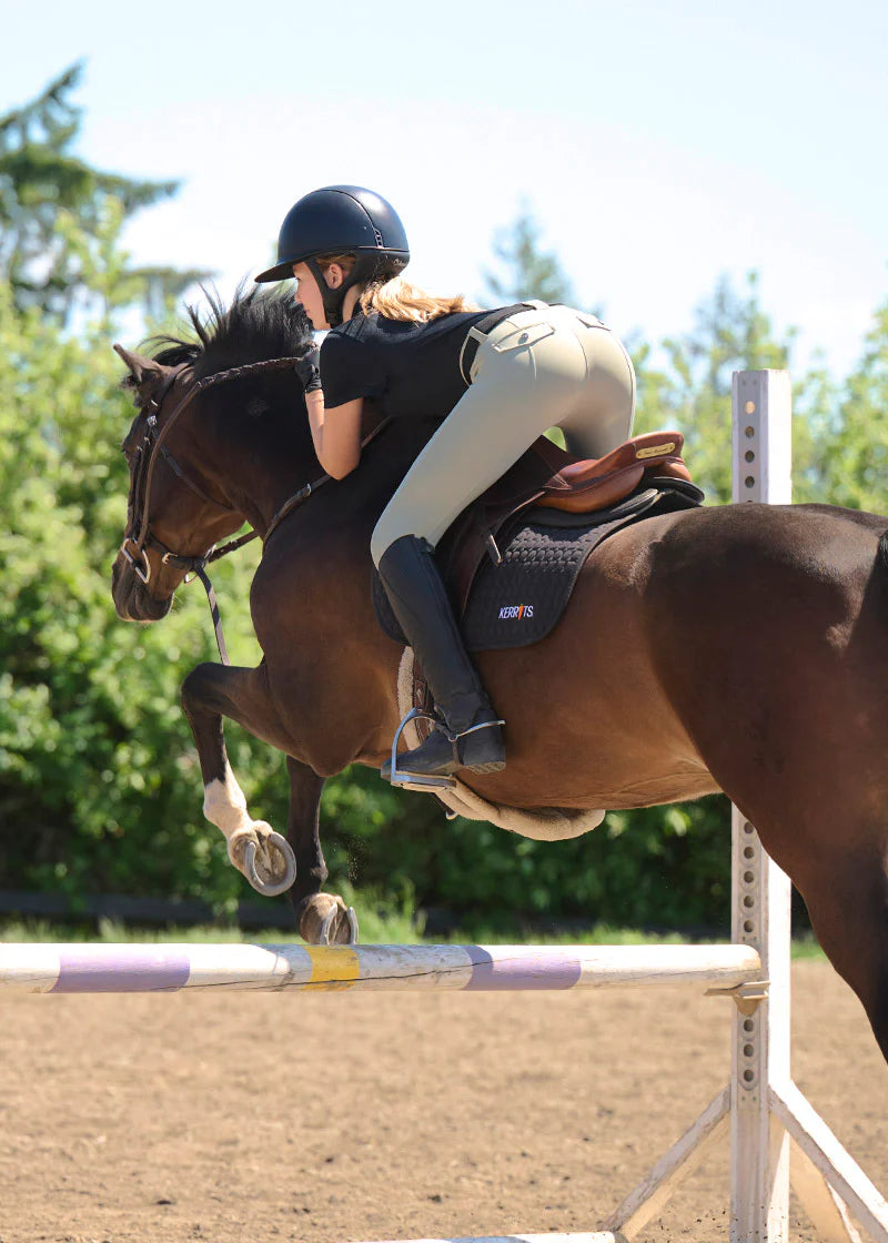 Back view of a young female model wearing the tan Kerrits Kids Free Motion Coolcore Knee Patch Breech and black shirt while jumping a bay horse