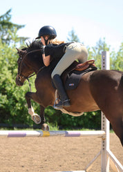 Back view of a young female model wearing the tan Kerrits Kids Free Motion Coolcore Knee Patch Breech and black shirt while jumping a bay horse