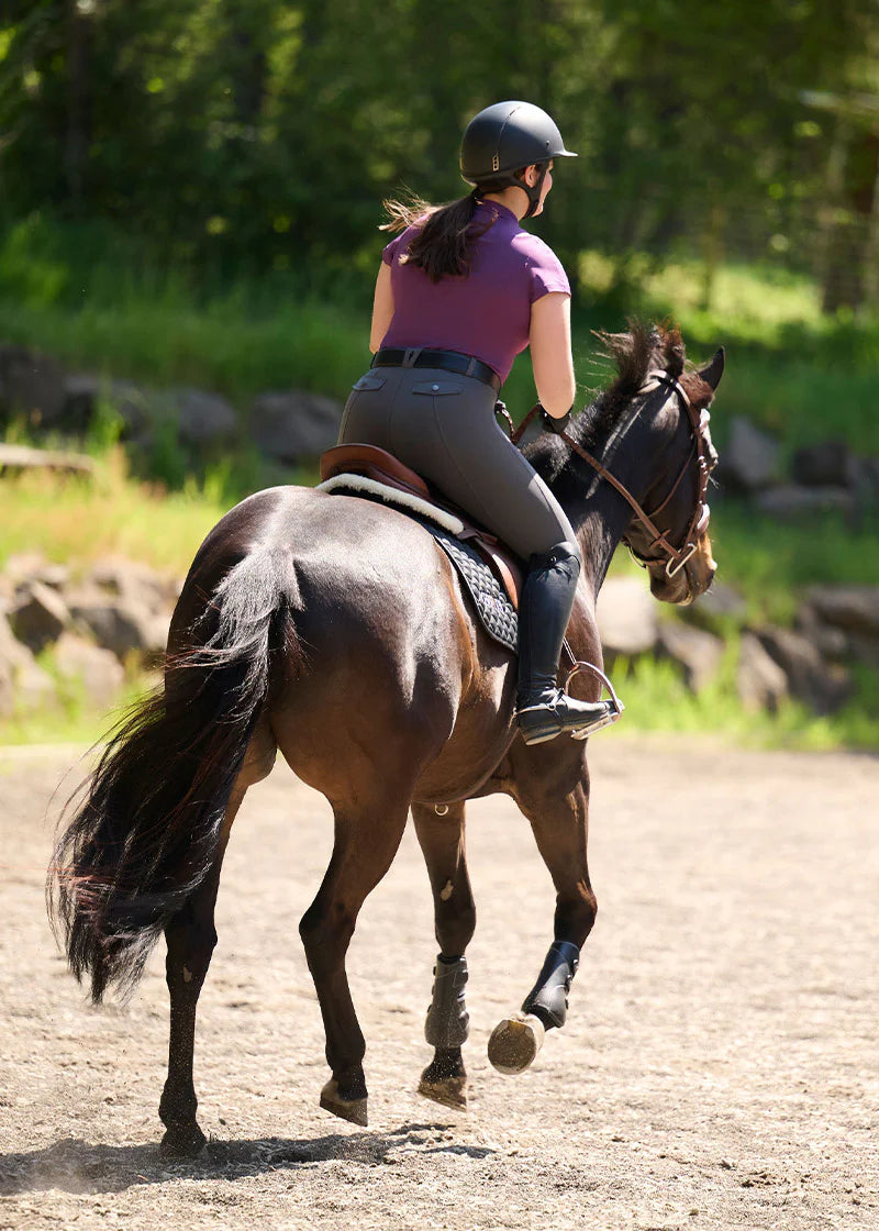 Back view of a female model wearing the purple Kerrits Serenity Coolcore Short Sleeve Polo while riding a dark colored horse