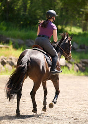 Back view of a female model wearing the purple Kerrits Serenity Coolcore Short Sleeve Polo while riding a dark colored horse