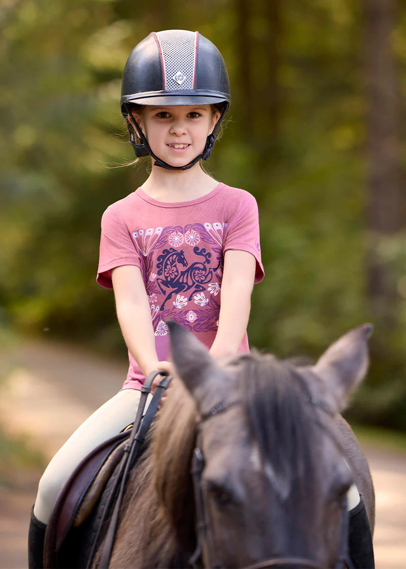 Front view of a young model wearing the pink Kerrits Kids Frolic Horse Tee while riding a horse outside