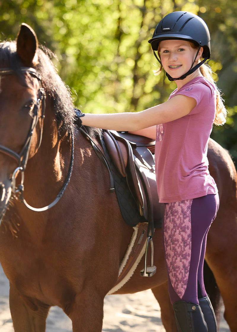 Side view of a young female model wearing the pink Kerrits Kids Frolic Horse Tee while standing next to a bay horse