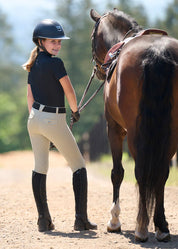 Back view of a young female model wearing the tan Kerrits Kids Free Motion Coolcore Knee Patch Breech and black shirt while standing next to a bay horse