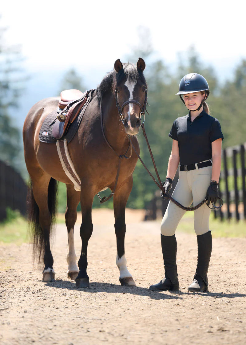 Full body front view of a young female model wearing the tan Kerrits Kids Free Motion Coolcore Knee Patch Breech and black shirt while standing next to a bay horse
