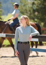 Front view of a female model wearing the light blue Kerrits Precision Long Sleeve Crew Neck Tech Top while leaning on a jump in an outdoor arena