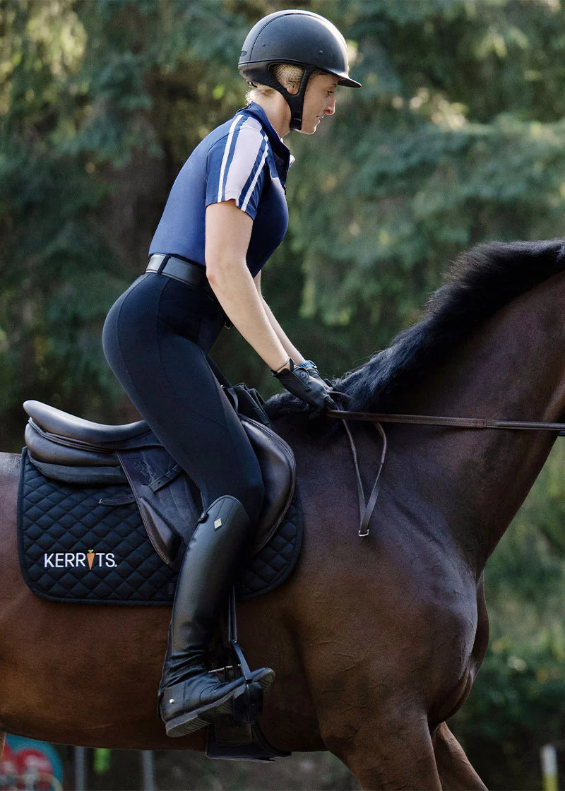 Side view of a female model wearing the black Kerrits Coolcore Full Leg Tech Tight while riding a horse