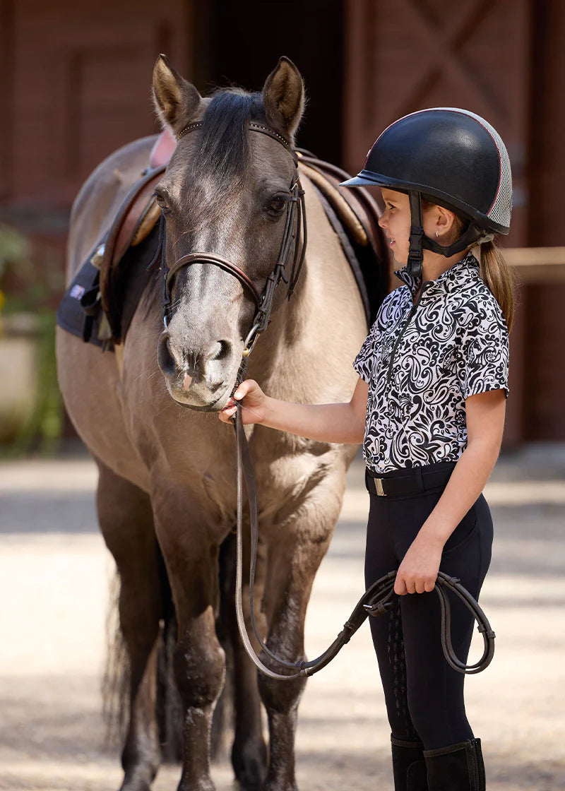 Side view of a young female model wearing the black Kerrits Kids Coolcore Full Seat Tech Tights and black and white patterned shirt while standing next to a horse