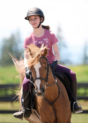 Front view of a young female model wearing the purple Kerrits Kids Performance Knee Patch Pocket Riding Tight while riding a chestnut horse outside