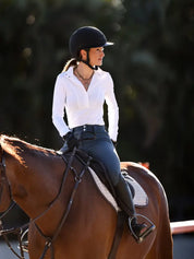 Front view of a female model wearing the white Criniere Helen Show Shirt while riding a bay horse