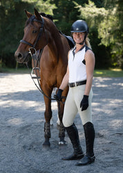 Full body front view of a female model wearing the tan Kerrits Affinity Pro Knee Patch Breech and white tank top while standing in front of a bay horse