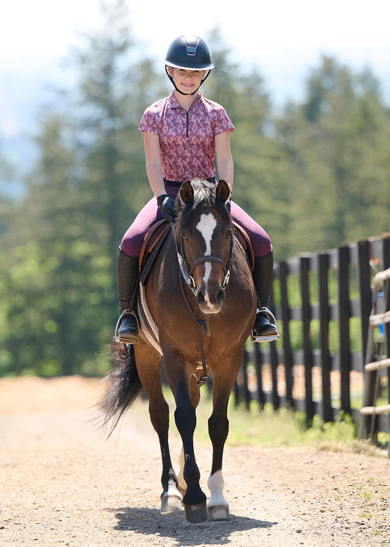 Fonrt view of a young female model wearing the pink Kerrits Kids EquiBreeze Ice Fil Short Sleeve Print Shirt while riding a brown horse in an outdoor arena