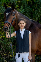 Model wearin the Helite Zip'In 2 Airbag in black with white breeches standing next to a bay horse