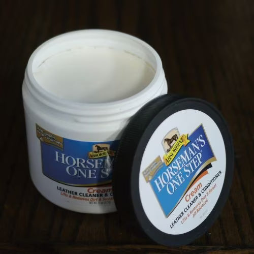 Front view of the Absorbine Horseman's One Step Cream Leather Cleaner & Conditioner with lid off and leaning up against the container on a wood background