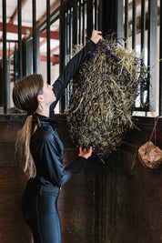 View of a female model hanging a black and purple Equi-Essentials Niblet Slow Feed Hay Net with hay in a stall