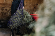 View of someone filling the black and purple Equi-Essentials Niblet Slow Feed Hay Net with hay while standing in a barn