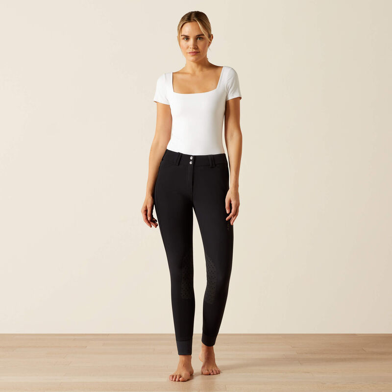Full body front view of a female model wearing the black Ariat Tri Factor Grip Knee Patch Breech and white shirt in a studio setting