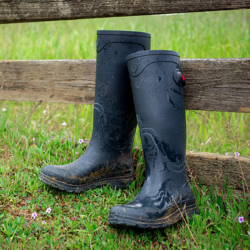 View of a pair of Navy Ariat Kelmarsh Rain Boot leaned up against a wood fence in the grass