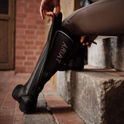 Rear view of a model sitting on a step and putting on a Ariat Heritage Contour Half Chap over a paddock boot
