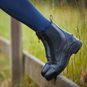 Side view of someone's feet propped up on an outdoor fence board and wearing the black Ariat Heritage Zip Paddock Boots