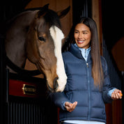Front view of a female model wearing the Big Dipper Ariat Ultrapuff Down Jacket while standing in a barn next to a horse in a stall