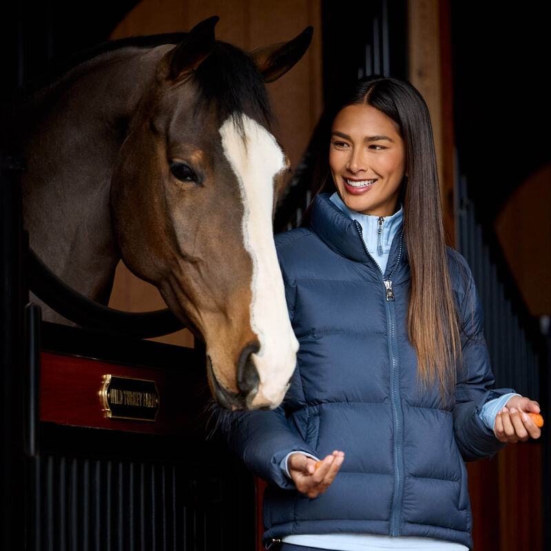 Front view of a female model wearing the Big Dipper Ariat Ultrapuff Down Jacket while standing in a barn next to a horse in a stall