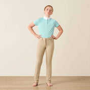 Full body front view of a young girl wearing a blue shirt and the Ariat Youth Protege Jod Knee Patch Breeches in tan in a studio setting