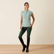 Full body front view of a model wearing the blue surf Ariat Breathe 1/2 Zip Baselayer, green tights, and black tall boots on a plain background