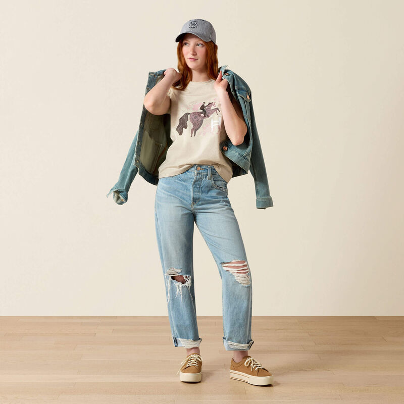 Full body front view of a young girl wearing the Ariat Youth Floral Leap T-Shirt in tan with jeans in a studio setting