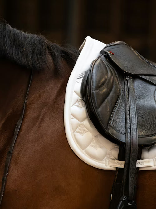 Side view of a bay horse wearing the white PS of Sweden Heart Jump Pad under a brown saddle