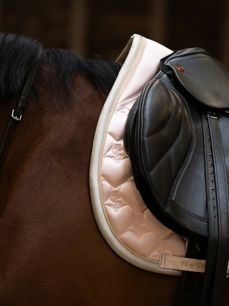 Side view of a bay horse wearing the light pink PS of Sweden Heart Jump Pad under a brown saddle