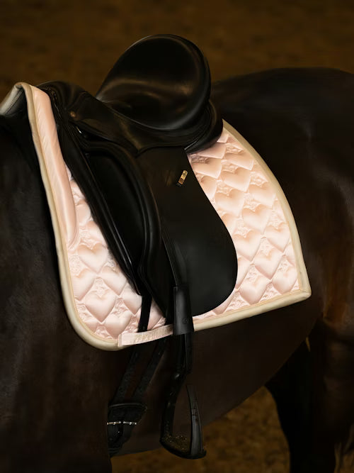 Angled side view of a black horse wearing the light pink PS of Sweden Heart Dressage Saddle Pad under a black saddle