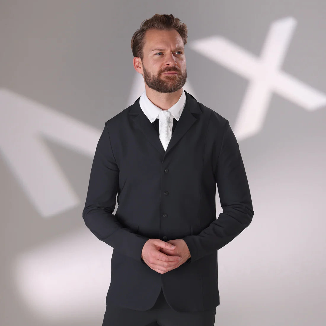 Front view of a male model wearing the black Aubrion Men's Optima MX Show Coat with the background being a blurry white MX on a gray