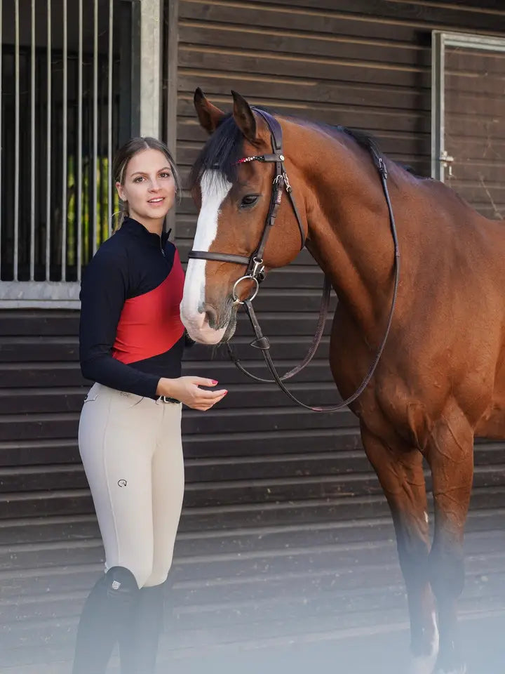 Angled front view of a bay horse and a female model who is wearing the black/red Criniere Sophie Fierce Focus Long Sleeve Shirt with a barn in the background