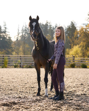 Side view of a young model wearing the Elderberry/Elderberry Fillies Kerrits Kids Thermo Tech Full Leg Tight and coordinating top while standing next to a horse in an outdoor arena