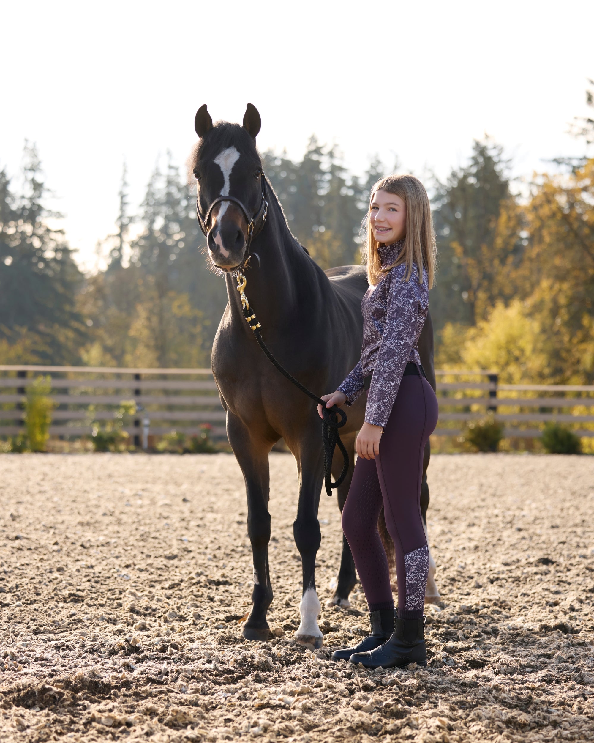 Side view of a young model wearing the Elderberry/Elderberry Fillies Kerrits Kids Thermo Tech Full Leg Tight and coordinating top while standing next to a horse in an outdoor arena
