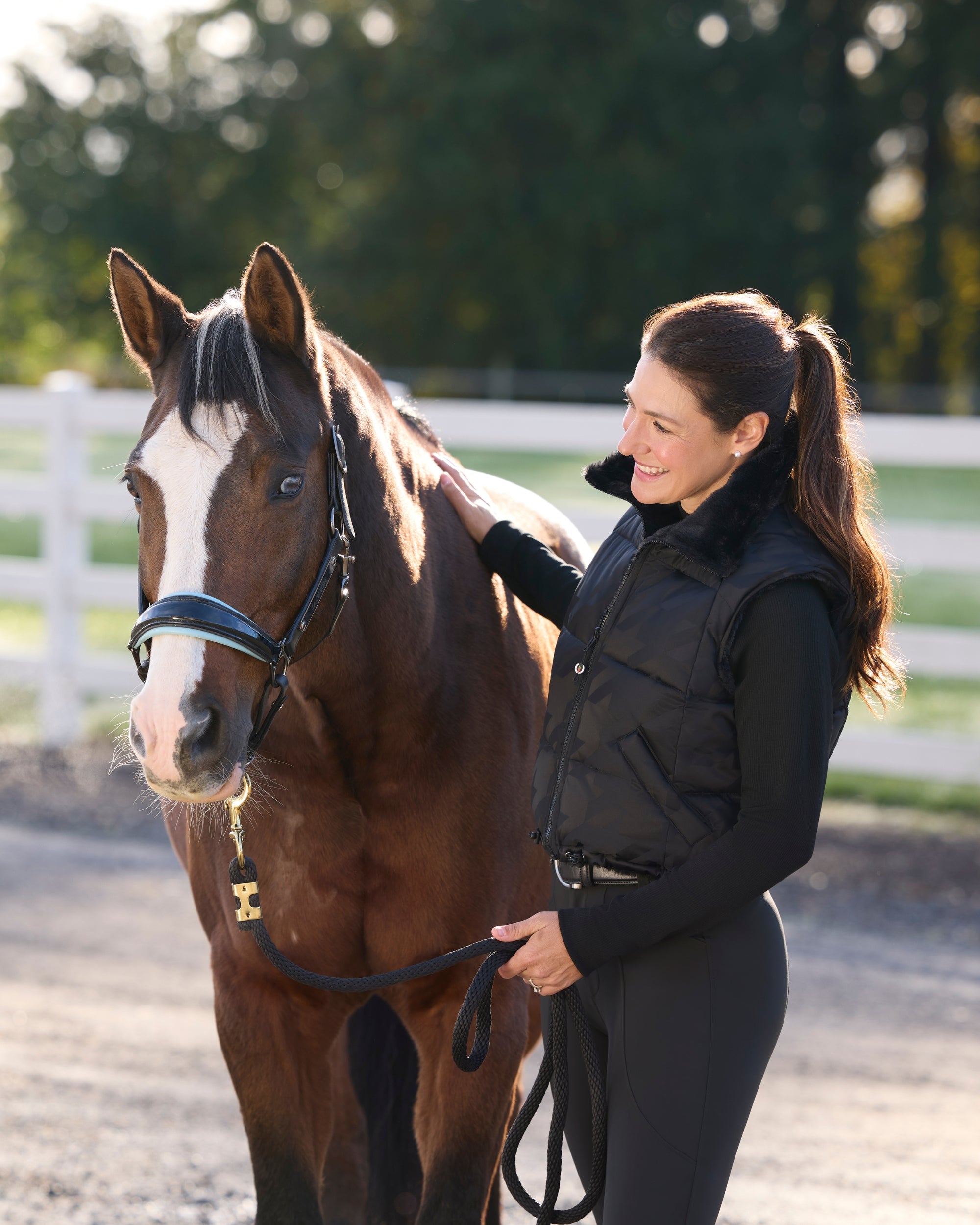 Angled front view of a female model wearing the Black Kerrits Align Textured Mock Neck under a black vest while standing next to a horse in an outdoor arena