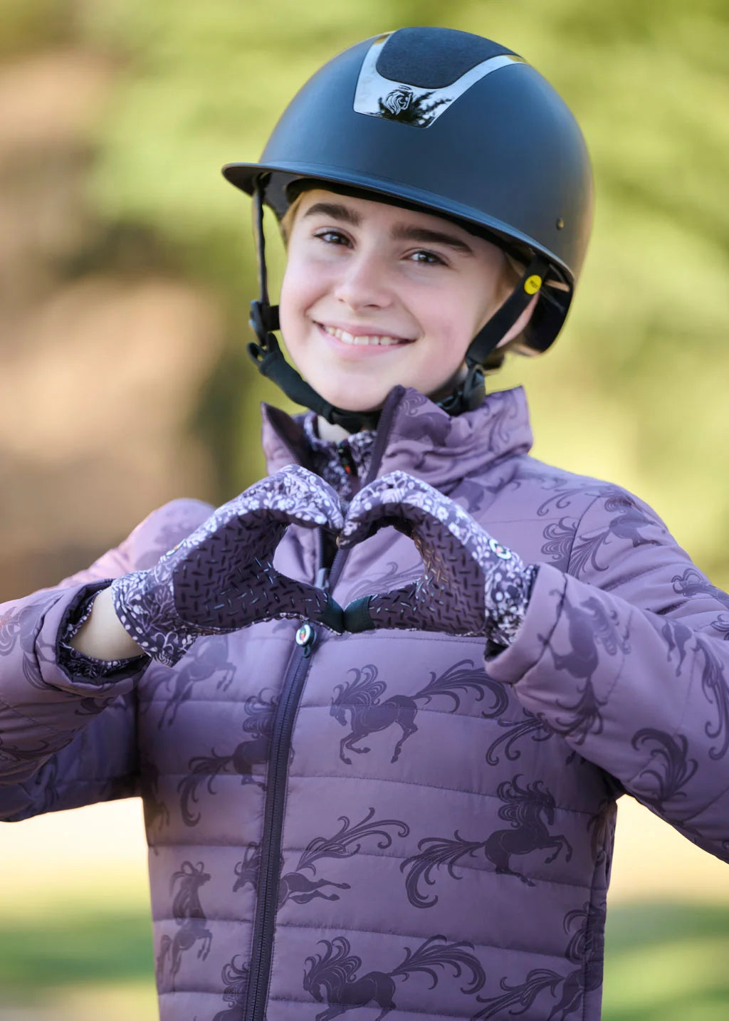 View of a young female model wearing the Elderberry Fantasy FIllies Kerrits Kids Thermo Tech Riding Gloves and coordinating jacket while making a heart shape with her hands