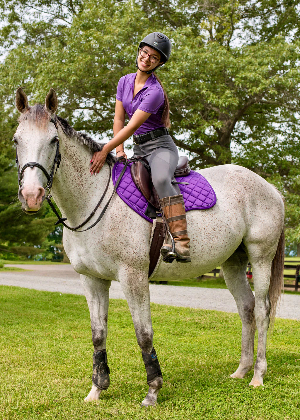 Side view of a female model ridingray horse who is wearing the Purple Kerrits Close Contact Saddle Pad while standing in the grass