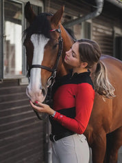 Front view of a bay horse and a female model who is wearing the black/red Criniere Sophie Fierce Focus Long Sleeve Shirt with a barn in the background