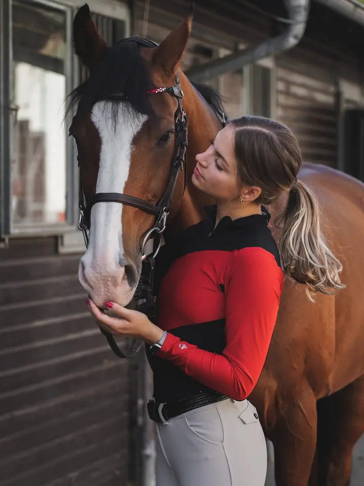Front view of a bay horse and a female model who is wearing the black/red Criniere Sophie Fierce Focus Long Sleeve Shirt with a barn in the background