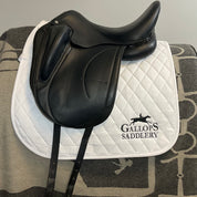 A black leather equestrian saddle resting on a white quilted saddle pad with the brand name 'Gallops Saddlery' printed in black. The saddle pad is laid on a gray and black patterned blanket against a plain light gray wall.