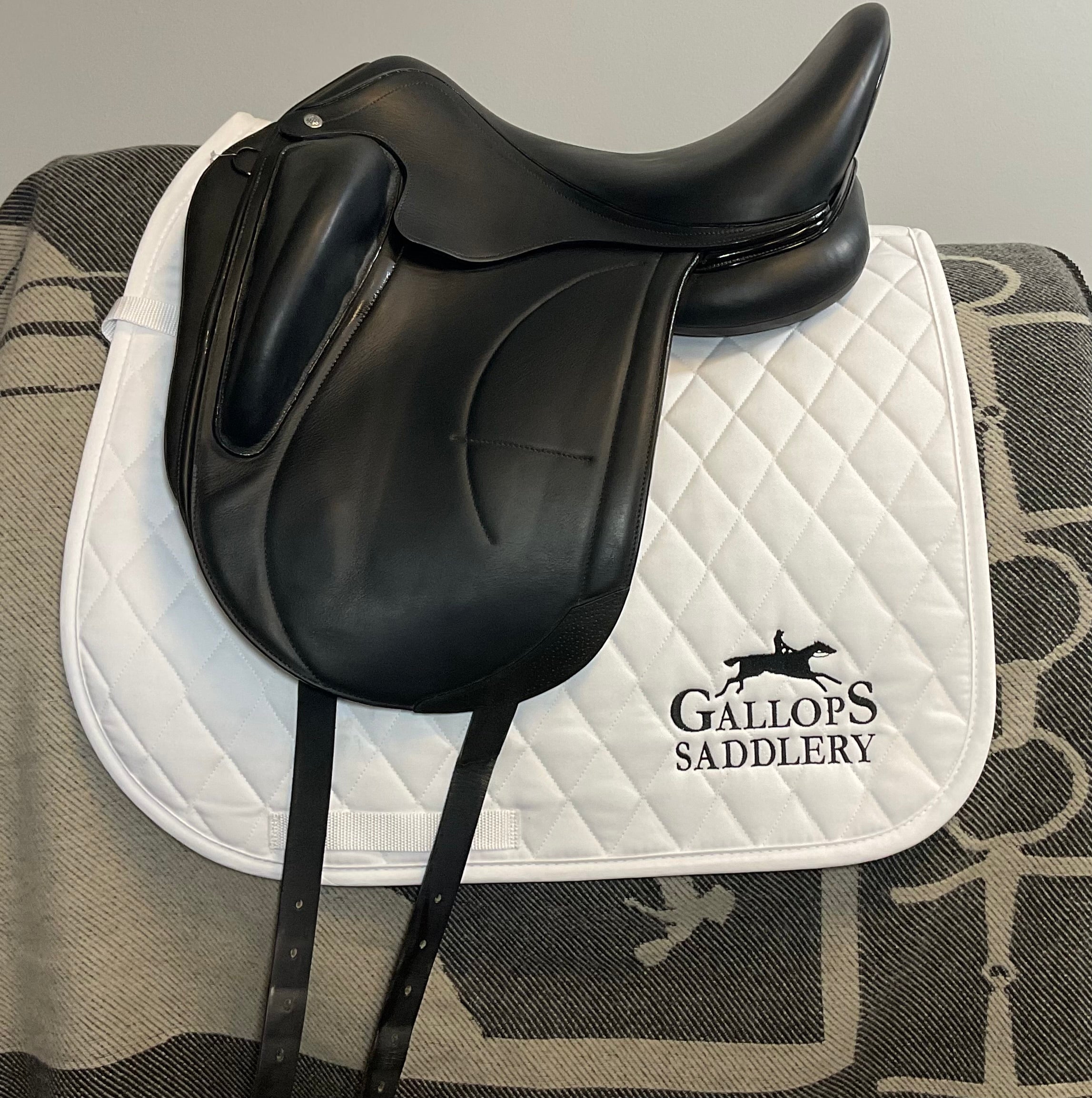 A black leather equestrian saddle resting on a white quilted saddle pad with the brand name 'Gallops Saddlery' printed in black. The saddle pad is laid on a gray and black patterned blanket against a plain light gray wall.