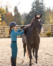 Side view of a young female model wearing the Jasper/Jasper Horseshoe Kerrits Kids Thermo Tech Full Leg Tight while standing next to a horse and adjusting her stirrups in an outdoor arena