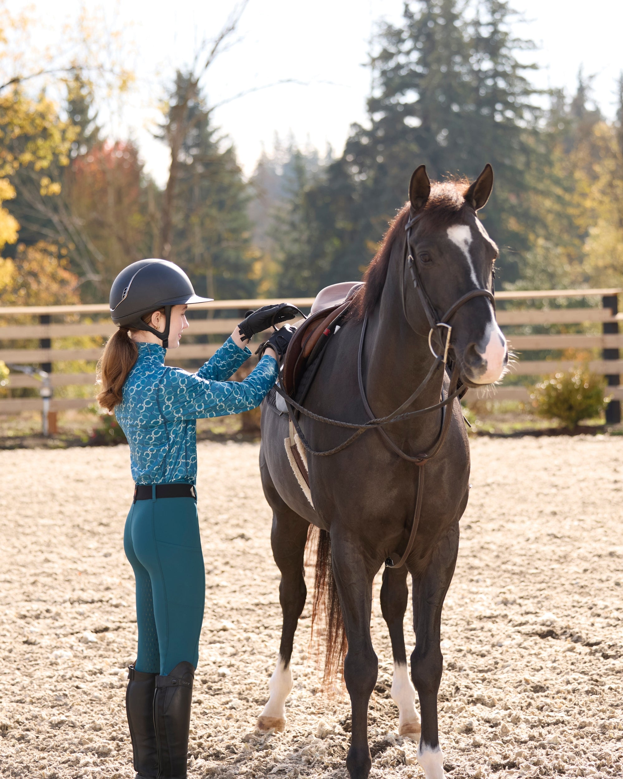 Side view of a young female model wearing the Jasper/Jasper Horseshoe Kerrits Kids Thermo Tech Full Leg Tight while standing next to a horse and adjusting her stirrups in an outdoor arena