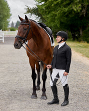 Side view of a female model wearing the white Kerrits Crossover II Full Seat Breech with black show coat while standing next to a bay horse on a gravel path