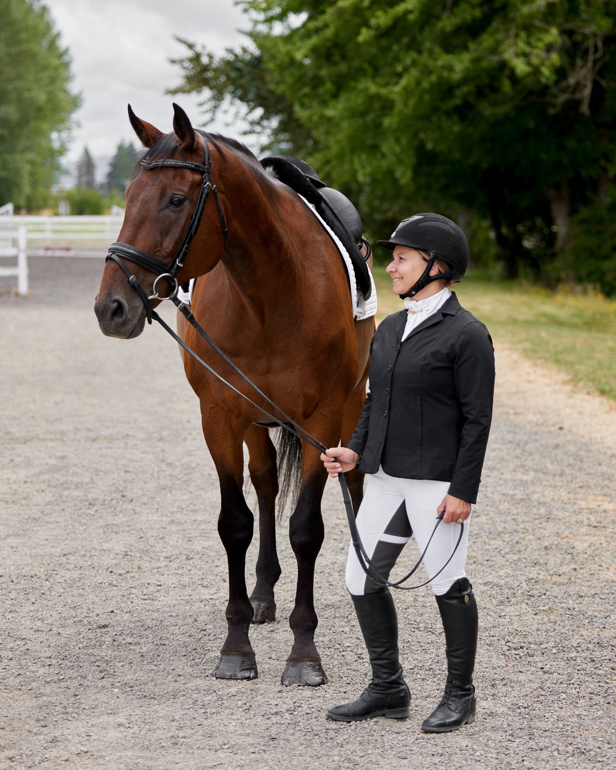 Side view of a female model wearing the white Kerrits Crossover II Full Seat Breech with black show coat while standing next to a bay horse on a gravel path