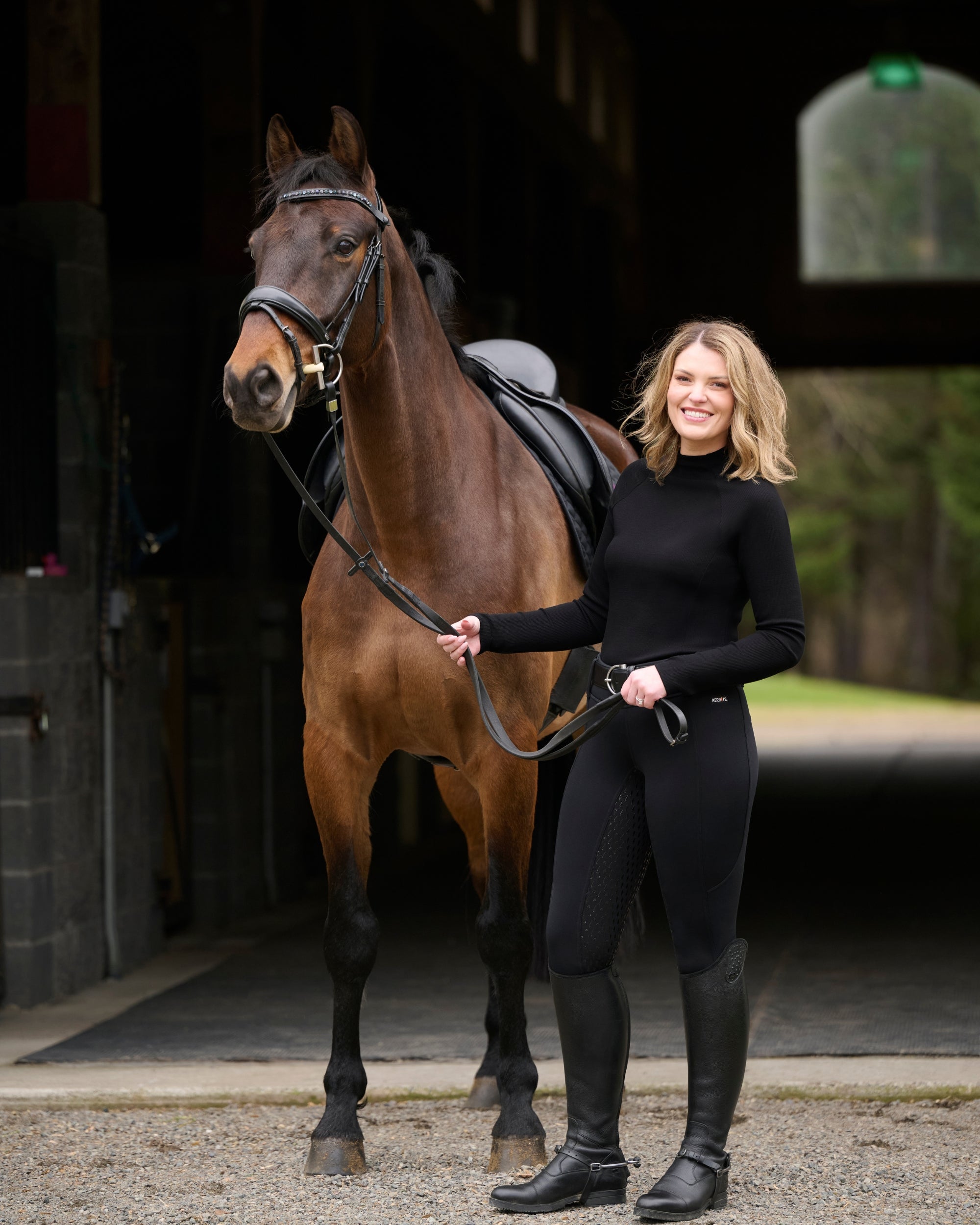 Front view of a female model wearing the Black Kerrits Align Textured Mock Neck while standing next to a bay horse in front of a barn aisle