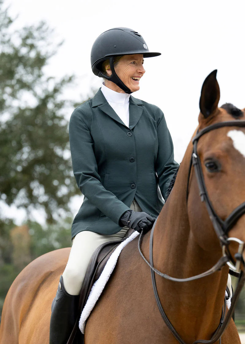 Front view of a female model wearing the green Kerrits Stretch Competitor Koat 3 Snap while riding a bay horse