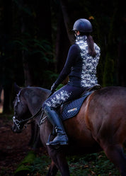 Front view of a model wearing the Black Happy Herds Kerrits Horse Reflection Quilted Vest while riding a horse on a trail in the woods